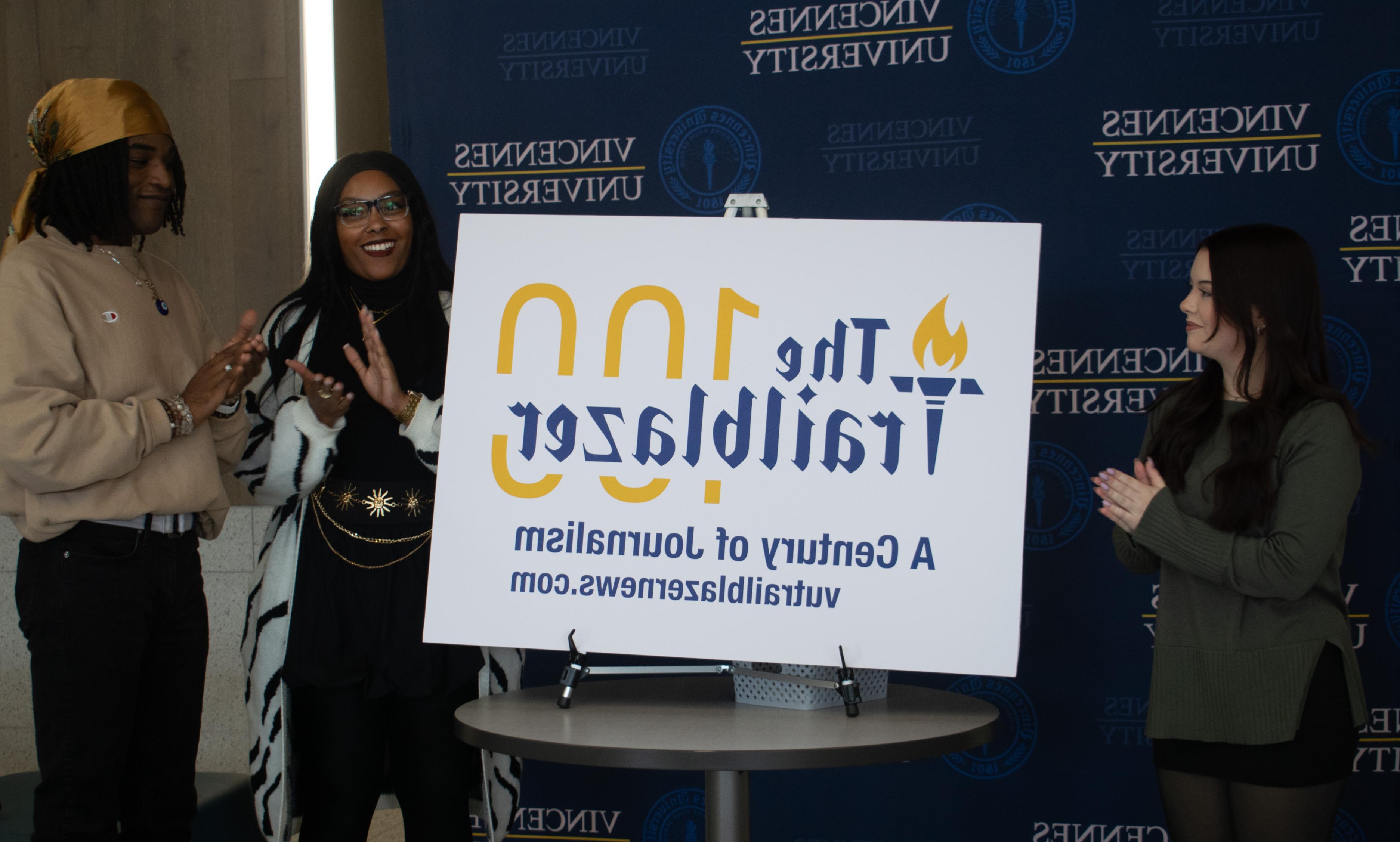 Three diverse college students including 2 female and 1 male clapping. Between them is a sign on a tabletop easle with the new logo for The Trailblazer newspaper. The logo says, The Trailblazer 100, a Century of Journalism and vutrailblazernews.com.